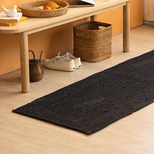 May include: A long, rectangular black jute rug. The rug is placed on a light wood floor. A wooden bench, a woven basket, and a pair of white sneakers are visible in the background, suggesting a home decor setting.