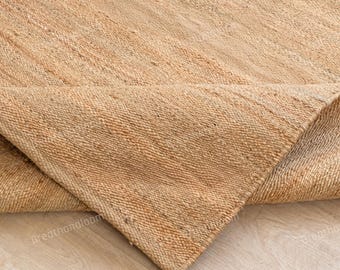 May include: Close-up of a natural jute rug with a folded corner. The rug has a woven texture and a light brown color. The corner reveals the rug's thickness and construction, showcasing its durable design. The rug is suitable for indoor use.