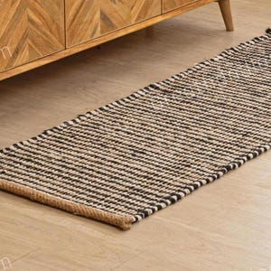 May include: A rectangular runner rug with a striped pattern in black and natural tones. The rug is made of woven material and has a brown border. The rug is placed on a light-colored wooden floor.