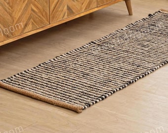 May include: A rectangular runner rug with a striped pattern in black and natural tones. The rug is made of woven material and has a brown border. The rug is placed on a light-colored wooden floor.
