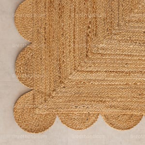 May include: A natural jute rug with a scalloped edge. The rug is a light brown colour and has a woven texture. The text "Greatthandloom" is printed on the rug.