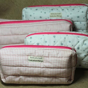 Peut inclure: Ensemble de quatre trousses de maquillage matelassées de différentes tailles. Deux sacs présentent un motif vichy rose et blanc, tandis que les deux autres ont un fond blanc avec des motifs d'étoiles grises. Chaque sac a une fermeture éclair rose vif et une étiquette "love yourself love your things."