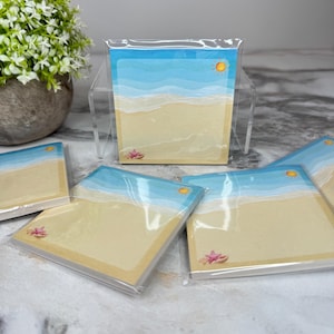 May include: Square notepads with a beach scene design. Each notepad features a blue sky, a yellow sun, and a sandy beach with a starfish. The notepads are packaged in clear plastic.