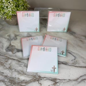 May include: Five square sticky note pads in clear plastic packaging. Each pad features the text GOD IS GOOD all the time in colorful letters at the top, with a small floral cross and a hand-drawn heart icon at the bottom right corner.