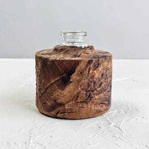 Puede incluir: Una botella decorativa de madera con una tapa de vidrio transparente. La botella es cilíndrica con una superficie rugosa y texturizada en tonos marrones. La tapa de vidrio tiene una pequeña abertura redondeada. La botella está sobre una superficie blanca.