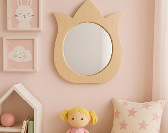 May include: A light wood-framed mirror in the shape of a tulip is mounted on a pink wall, alongside framed bunny and cloud art. A doll in a pink polka-dot dress and a star-patterned pillow sit below the mirror, creating a nursery theme.