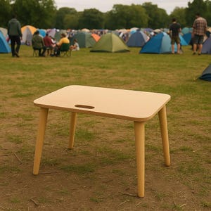 May include: A small, light-colored wooden table with rounded corners and four angled legs. The tabletop has a handle cutout. The table is set up on a grassy field, with tents and people in the background, suggesting outdoor use.