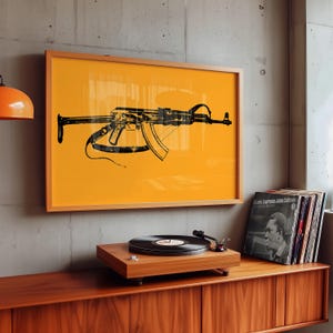 May include: A rectangular framed wall art piece featuring a black silhouette illustration of an AK-47 rifle centred on a solid vibrant orange background. The artwork is displayed in a light wood frame, mounted on a grey concrete wall above a wooden cabinet.