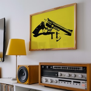 Könnte beinhalten: Ein gerahmter Wandkunstdruck mit einer schwarzen Silhouettenillustration eines Revolvers und drei Patronen vor einem einfarbigen gelben Hintergrund, eingefasst in einen hellen Holzrahmen an einer weißen Wand.