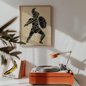 May include: A framed wall art print featuring a black silhouette of a gladiator holding a sword and shield on a beige background. Below, an orange record player sits next to a vinyl record sleeve displaying a yellow banana and the text Andy Warhol.