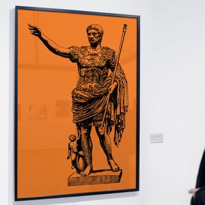 May include: A framed wall art print featuring a black ink illustration of a Roman emperor in military armour, standing with one arm outstretched and holding a staff, set against a solid, vibrant orange background within a thin black frame.