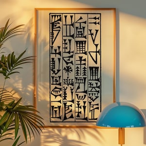 May include: A framed art print featuring black abstract characters on a white background. The artwork is encased in a thin, gold-colored frame. The image is displayed on a white wall, with a blue lamp and a green plant in the foreground.