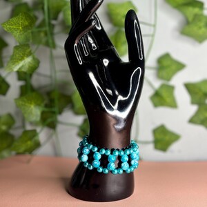 May include: A turquoise beaded bracelet displayed on a black hand-shaped jewelry holder. The bracelet features multiple strands of round, light blue beads. The background includes green leafy vines and a pink surface.