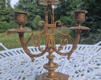 Puede incluir: Candelabro dorado ornamentado con tres candeleros. El candelabro tiene una base decorativa y brazos curvos. El diseño incluye detalles florales y de volutas. El fondo muestra una mesa blanca y árboles verdes.