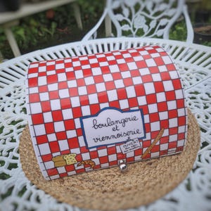 Könnte beinhalten: Eine rechteckige Brotdose mit rot-weißem Karomuster. Auf der Dose steht in einem blauen Rahmen "boulangerie et viennoiserie", sowie Abbildungen von Brot und einem Croissant. Ein kleines Schild zeigt "Ouvert du soir au matin".