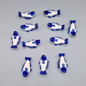 Peut inclure: Onze figurines de poissons en céramique, blanches avec des détails bleus sur la tête, la queue et les nageoires. Les figurines sont disposées sur une surface grise, mettant en valeur leur aspect décoratif. Chaque poisson mesure environ 7 cm de long.
