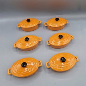 Könnte beinhalten: Sechs ovale, orangefarbene Mini-Cocottes aus Keramik mit schwarzen Knaufdeckeln und seitlichen Griffen. Die Cocottes sind auf einer hellgrauen Oberfläche angeordnet und zeigen ihre leuchtende Farbe und ihr Design. Ideal für Einzelportionen.