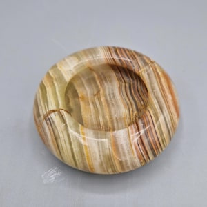 May include: A round ashtray made of natural stone, featuring a mix of beige, brown, and green tones. The ashtray has a circular design with a central depression for holding ashes and cigarettes. The stone has a polished finish.