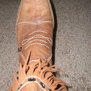 May include: A tan leather boot with a pointed toe and decorative white stitching. The boot features a fringe detail around the ankle and is adorned with silver-toned metal studs. The boot is a size 8.