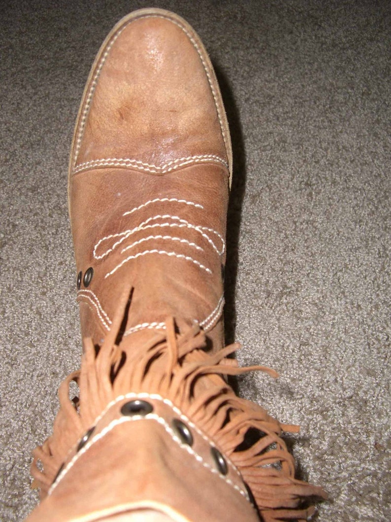 May include: A tan leather boot with a pointed toe and decorative white stitching. The boot features a fringe detail around the ankle and is adorned with silver-toned metal studs. The boot is a size 8.