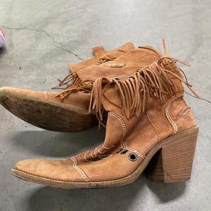 May include: A pair of brown suede cowboy boots with fringe detailing. The boots have a pointed toe, a block heel, and white stitching accents. The boots are worn and have a vintage look.