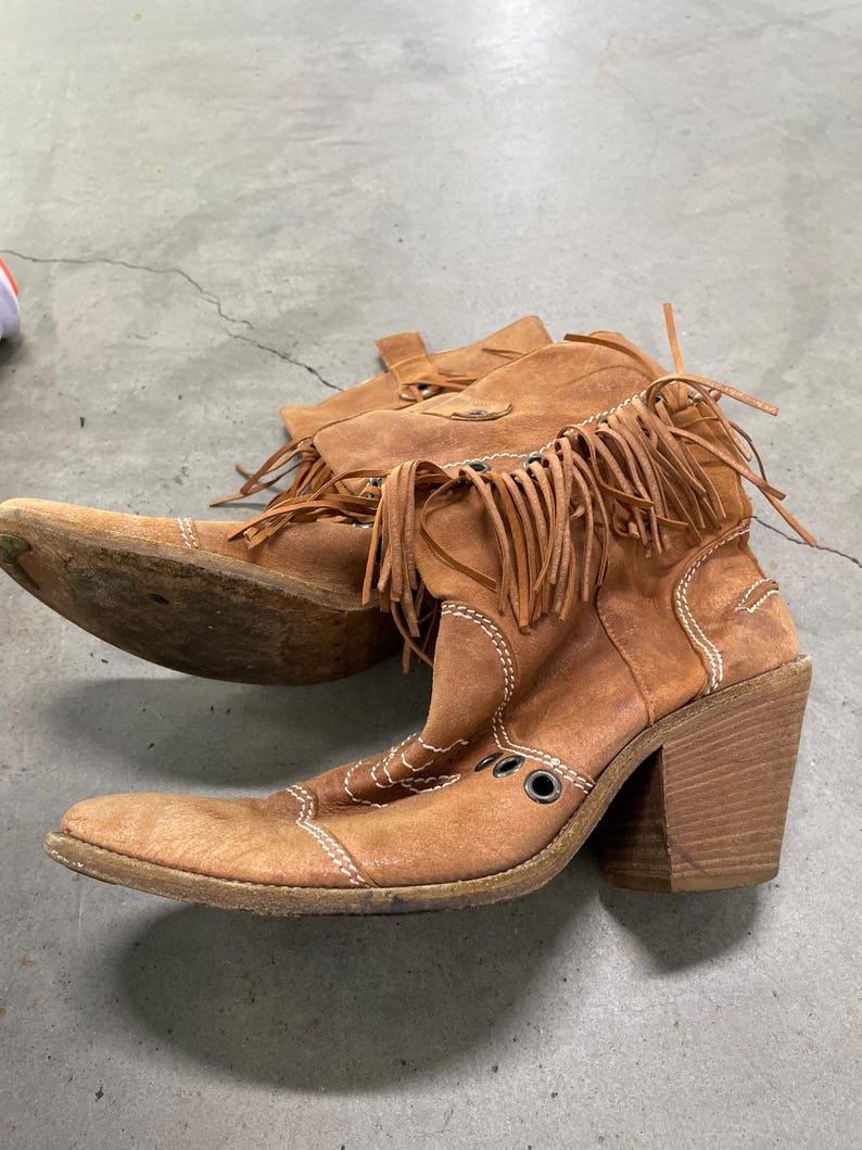 May include: A pair of brown suede cowboy boots with fringe detailing. The boots have a pointed toe, a block heel, and white stitching accents. The boots are worn and have a vintage look.