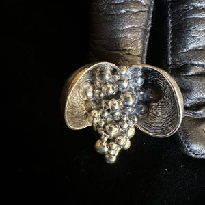 Op de afbeelding: Een zilverkleurige ring met een cluster van kleine, ronde kralen in het midden. De ring heeft een uniek, open ontwerp met twee gebogen, bloembladachtige vormen aan weerszijden van de kralencluster. De ring is geplaatst tegen een zwarte achtergrond en zwart leer.