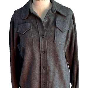 May include: A charcoal gray button-down shirt with two chest pockets and a collared neckline. The long-sleeved shirt is displayed on a mannequin. The fabric appears to be a soft, wool blend. The shirt has a classic, vintage style.