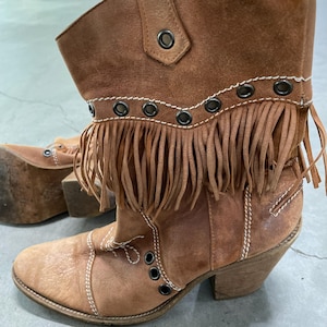 May include: Brown leather ankle boots with fringe detailing and metal grommets. The boots have a stacked heel and decorative stitching. The boots are a light brown color and have a worn appearance.