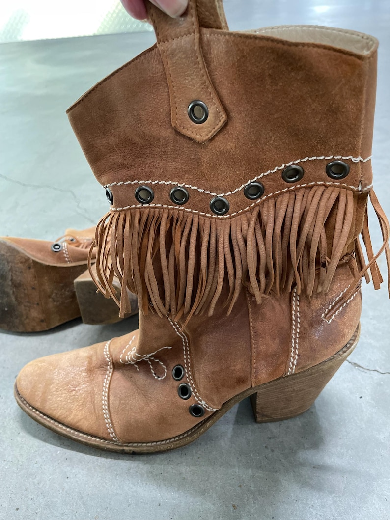 May include: Brown leather ankle boots with fringe detailing and metal grommets. The boots have a stacked heel and decorative stitching. The boots are a light brown color and have a worn appearance.