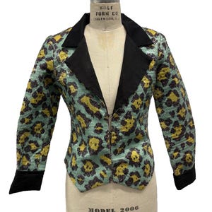 May include: A patterned jacket on a mannequin. The jacket features a teal background with yellow and black leopard print. It has a black collar, cuffs, and a zipper closure. The mannequin is labeled "WOLF FORM CO." and "MODEL 2006".