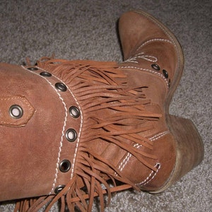 May include: Brown suede cowboy boot with fringe detail. The boot features a pointed toe, a low heel, and decorative stitching. Metal grommets accent the boot's design. A stylish Western-inspired footwear option.