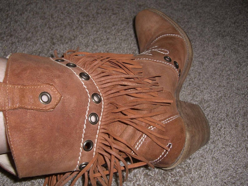 May include: Brown suede cowboy boot with fringe detail. The boot features a pointed toe, a low heel, and decorative stitching. Metal grommets accent the boot's design. A stylish Western-inspired footwear option.