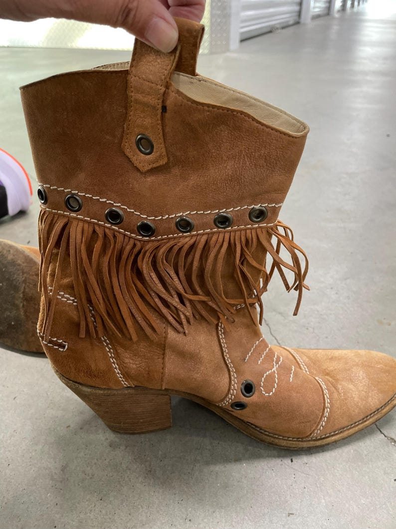 May include: A brown suede ankle boot with a pointed toe and a stacked heel. The boot features fringe detailing along the top and decorative stitching. Metal grommets are visible along the top edge. The boot has a pull tab.