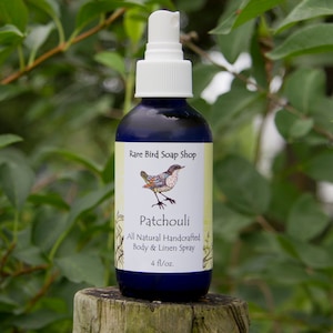 Puede incluir: Una botella de vidrio azul oscuro con un pulverizador blanco, etiquetada como "Patchouli" de Rare Bird Soap Shop. La etiqueta presenta una ilustración de un pájaro y dice "All Natural Handcrafted Body & Linen Spray", con una capacidad de 118 ml.