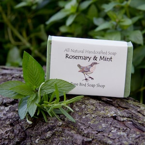 May include: A bar of natural handcrafted soap with a green and white label. The label reads "All Natural Handcrafted Soap Rosemary & Mint" and "Rare Bird Soap Shop". The soap is sitting on a tree branch with green leaves.