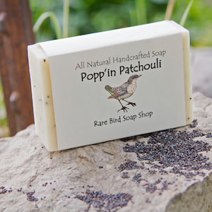 Puede incluir: Una pastilla de jabón artesanal con una etiqueta blanca que dice "All Natural Handcrafted Soap Popp'in Patchouli" y "Rare Bird Soap Shop". La etiqueta también presenta una ilustración colorida de un pájaro.