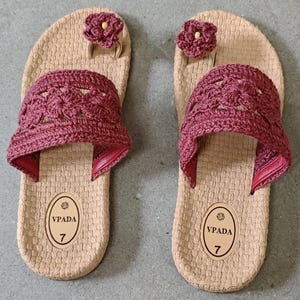 May include: A pair of beige flip-flops with a textured sole. Each sandal features a burgundy crochet strap and a burgundy flower accent. The sandals have the text "VPADA" and the number "7" printed on them.