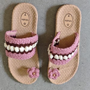 May include: A pair of beige sandals with pink and white crochet straps and a small pink flower accent. The sandals have a textured sole and the number 7 is printed on the insole.