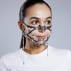 May include: A silver metal face mask with an elaborate, geometric design. The mask covers the nose, cheeks, and chin, with dangling chain details. The mask has a futuristic, avant-garde aesthetic.