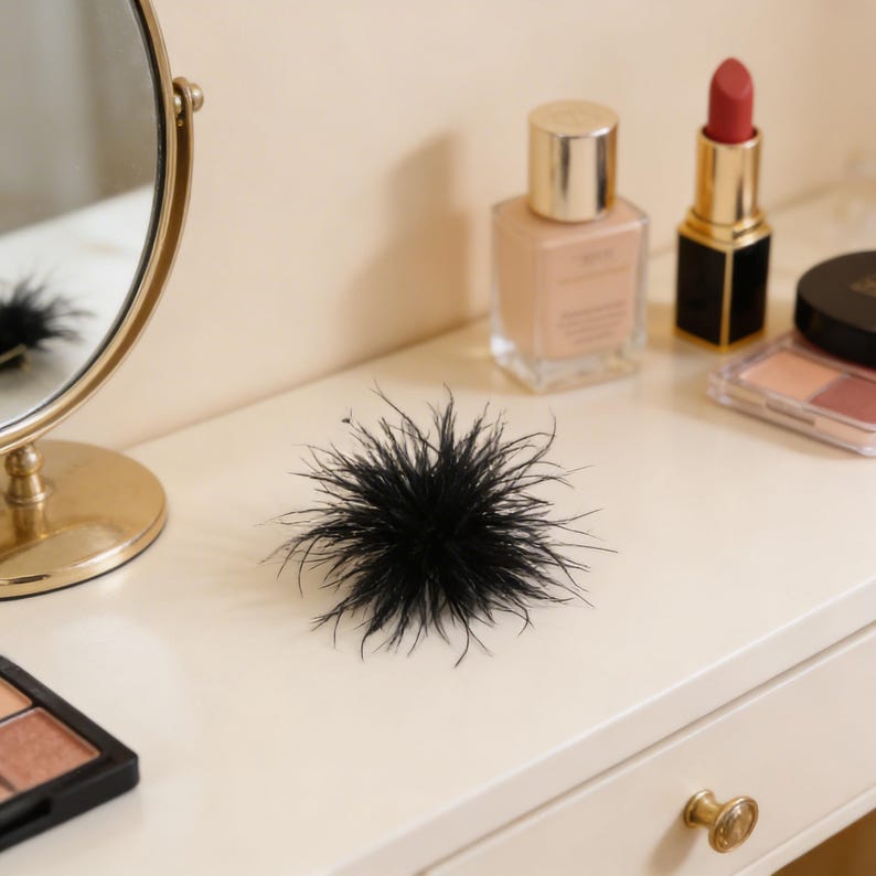 May include: A black feather hair accessory sits on a white vanity table. Makeup products, including foundation, lipstick, and eyeshadow, are arranged nearby. A gold-framed mirror is visible in the background, reflecting the scene.