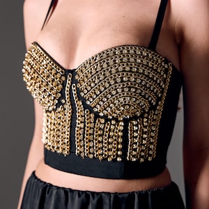 Puede incluir: Top bustier negro adornado con cadenas doradas y detalles brillantes. El top presenta un escote corazón y finas correas negras. Las cadenas caen por la parte delantera, creando un diseño glamuroso y llamativo.