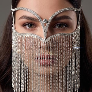 May include: A silver-toned face veil, adorned with numerous sparkling rhinestones, cascades down in vertical strands. The veil features a decorative headpiece with a central jewel and a curved design. The image is a close-up, highlighting the intricate details of the jewelry.