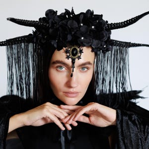 May include: A black headpiece with floral and horn-like details. The headpiece has a central jewel and dangling fringe. The person in the image is wearing a black outfit. The headpiece is an elaborate and gothic-inspired accessory.