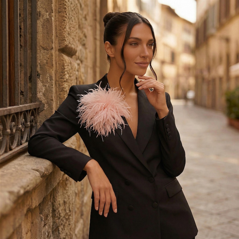 May include: A black double-breasted blazer with a light pink feather accent. The blazer has a notched lapel and button closures. The background shows a stone wall and a street.