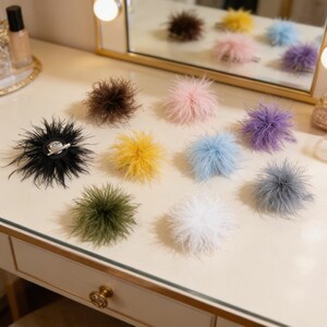 May include: A collection of colorful feather hair clips in various shades, including black, brown, pink, yellow, blue, green, white, and purple. Each clip features a round, fluffy feather design with a metal clip backing. The clips are arranged on a white surface.