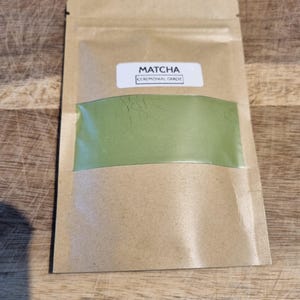 Pu&ograve; includere: Una busta di carta kraft contenente polvere di matcha. La busta ha una finestra trasparente che rivela la vibrante polvere verde. L'etichetta recita "Matcha Ceremonial Grade". La confezione &egrave; sigillata e pronta all'uso.