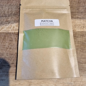 Pu&ograve; includere: Un sacchetto di carta marrone contenente polvere di matcha con una finestra trasparente che mostra la polvere verde. L'etichetta recita "Matcha Ceremonial Grade". Il sacchetto &egrave; sigillato e si trova su una superficie di legno.