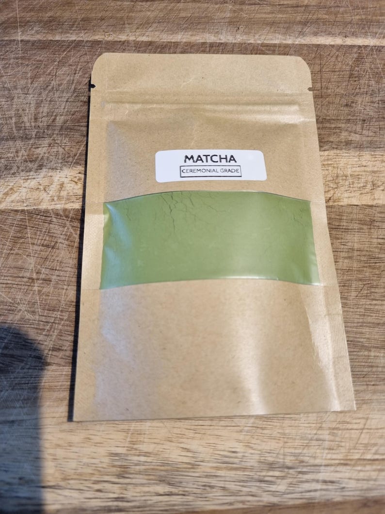 Pu&ograve; includere: Un sacchetto di carta marrone contenente polvere di matcha con una finestra trasparente che mostra la polvere verde. L'etichetta recita "Matcha Ceremonial Grade". Il sacchetto &egrave; sigillato e si trova su una superficie di legno.
