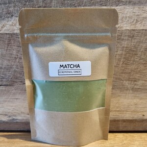 Pu&ograve; includere: Una busta di carta kraft con una finestra trasparente che mostra polvere di matcha verde. Un'etichetta bianca sulla parte anteriore recita "MATCHA CEREMONIAL GRADE". La busta &egrave; sigillata.